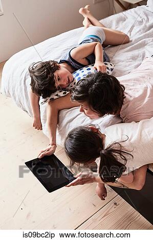 Stock Photography - Mother, son and daughter lying on bed, using digital tablet, elevated view. Fotosearch
