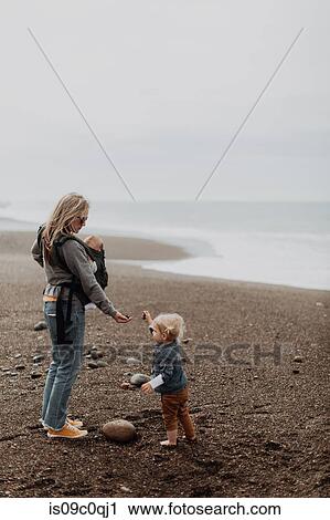Stock Image - Mother with baby in carrier playing with toddler on beach. Fotosearch