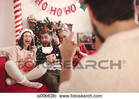 Stock Image - Over shoulder view of young man photographing friends on sofa at christmas party. Fotosearch