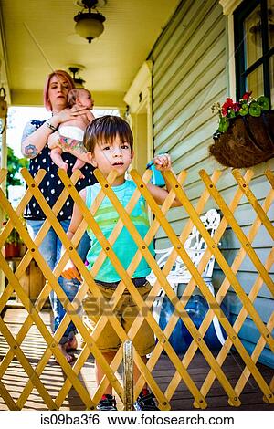 Stock Photograph - Portrait of male toddler with mother and baby sister by porch fence. Fotosearch