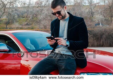 Picture - Portrait of man leaning against car texting on mobile phone. Fotosearch