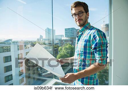 Stock Image - Portrait of man, standing beside office window, looking at plans. Fotosearch