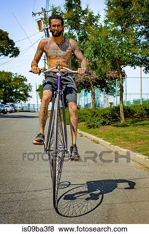Portrait of tattooed young man cycling suburban road on penny farthing View Large Photo Image Stock Photo - Portrait of tattooed young man cycling suburban road on penny farthing. Fotosearch