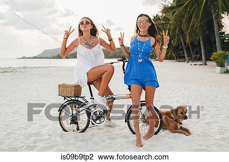 Stock Image - Portrait of two young women with bicycle making peace sign on sandy beach, Krabi, Thailand. Fotosearch