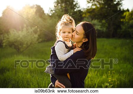Stock Image - Portrait of woman kissing her daughter in sunlit field. Fotosearch