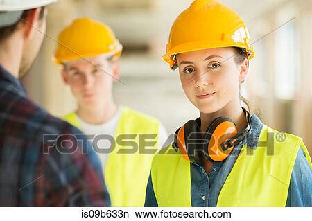 Picture - Portrait of woman wearing hard hat and hi vis vest, co-workers in background. Fotosearch