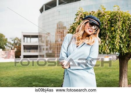 Stock Photograph - Portrait of woman wearing sunglasses and baker boy cap looking at camera smiling. Fotosearch
