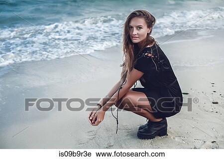 Stock Image - Portrait of young woman crouching by water's edge on beach, Odessa, Odessa Oblast, Ukraine. Fotosearch