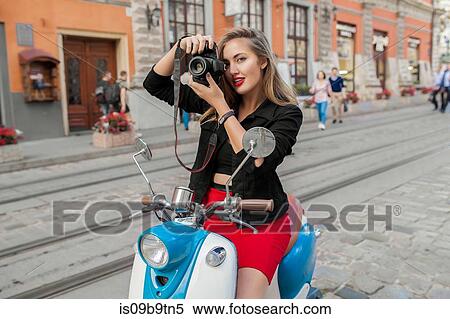 Stock Photography - Portrait of young woman on scooter taking photograph on cobbled street, Odessa, Ukraine. Fotosearch