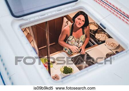 Stock Photography - Portrait of young woman through yacht hatch preparing food in kitchen, Croatia. Fotosearch
