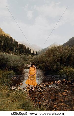 Stock Photography - Pregnant woman in orange maxi dress crossing stepping stones in rural river, Mineral King, California, USA. Fotosearch