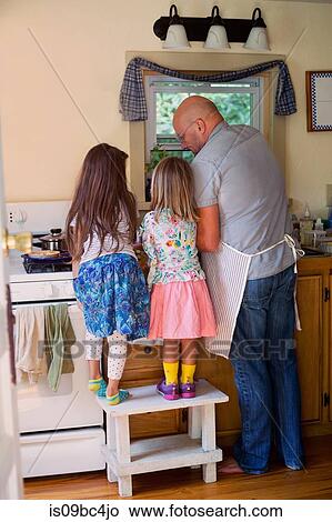 Rear view of girl and sister on stool watching father preparing food in kitchen View Large Photo Image Picture - Rear view of girl and sister on stool watching father preparing food in kitchen. Fotosearch
