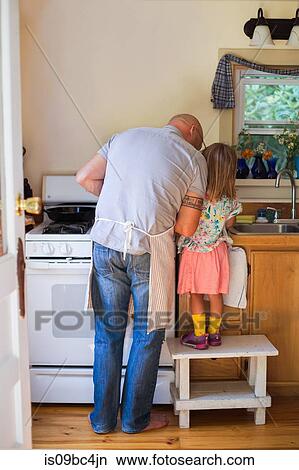 Picture - Rear view of girl on stool watching father preparing food in kitchen. Fotosearch
