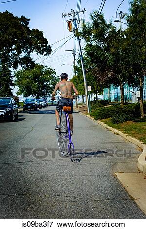 Stock Photo - Rear view of tattooed young man cycling suburban road on penny farthing. Fotosearch