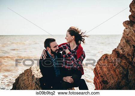 Stock Image - Romantic mid adult couple sitting on beach rock, Odessa Oblast, Ukraine. Fotosearch