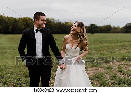 Stock Image - Romantic young bride and groom holding hands and laughing in field. Fotosearch