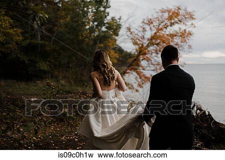 Romantic young groom carrying bride's wedding dress train on lakeside, rear view View Large Photo Image Stock Photo - Romantic young groom carrying bride's wedding dress train on lakeside, rear view. Fotosearch