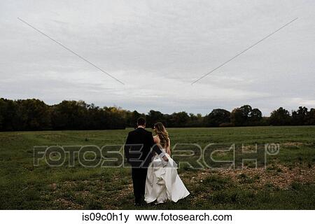 Stock Photo - Romantic young groom carrying bride's wedding dress train in field, rear view. Fotosearch