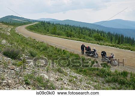 Stock Image - Senior male motorcyclist on rural mountain roadside with motorbike, high angle portrait, Dawson Creek, Canada. Fotosearch