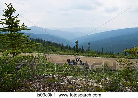 Stock Image - Senior male motorcyclist waving from rural mountain roadside with motorbike, high angle portrait, Dawson Creek, Canada. Fotosearch