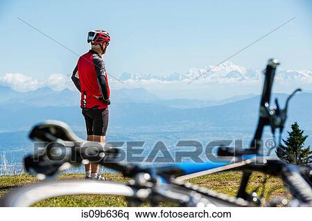 Stock Photography - Senior man wearing cycling helmet, standing on hillside, looking at view, bicycle in foreground. Fotosearch