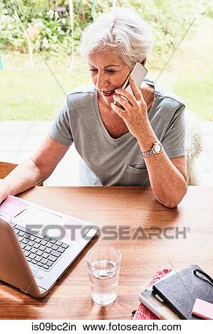 Senior woman sitting at table with laptop in front of her, using smartphone View Large Photo Image Picture - Senior woman sitting at table with laptop in front of her, using smartphone. Fotosearch