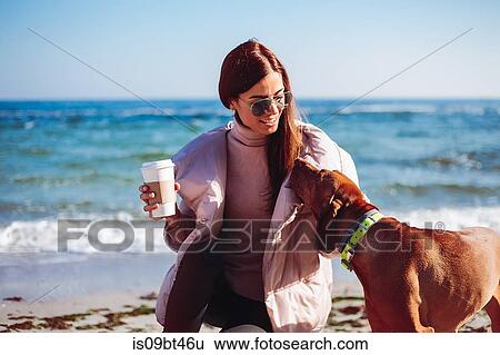 Stock Photograph - Stylish mid adult woman crouching on beach petting her dog, Odessa, Odeska Oblast, Ukraine. Fotosearch