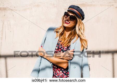 Stylish woman wearing sunglasses and cap in front of wall with arms folded View Large Photo Image Stock Image - Stylish woman wearing sunglasses and cap in front of wall with arms folded. Fotosearch
