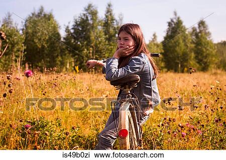 Stock Image - Teenage girl sitting on bicycle in long grassy field. Fotosearch