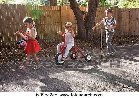 Stock Image - Three children in mini parade, banging drum, riding tricycle and using scooter. Fotosearch
