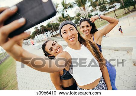 Stock Image - Three female friends, wearing sports clothing, taking selfie using smartphone, South Point Park, Miami Beach, Florida, USA. Fotosearch