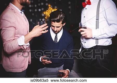 Stock Photography - Three men at party, man in middle using smartphone. Fotosearch