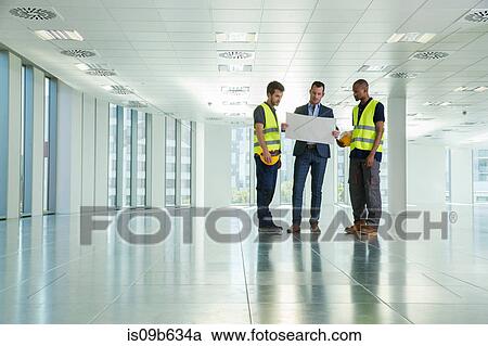 Stock Image - Three men standing in empty office, looking at plans. Fotosearch