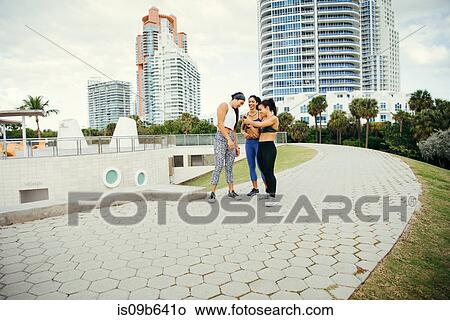 Three women wearing sports clothing, standing together, looking at smartphone, South Point Park, Miami Beach, Florida, USA View Large Photo Image Picture - Three women wearing sports clothing, standing together, looking at smartphone, South Point Park, Miami Beach, Florida, USA. Fotosearch