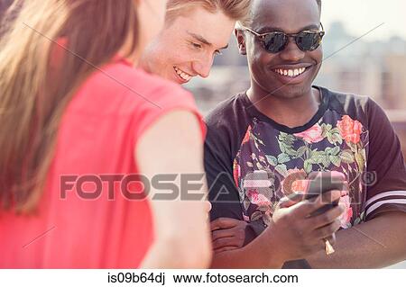 Stock Image - Three young adult friends looking at smartphone at roof party in London, UK. Fotosearch
