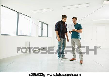 Stock Image - Two men standing in empty office space, looking at digital tablet. Fotosearch