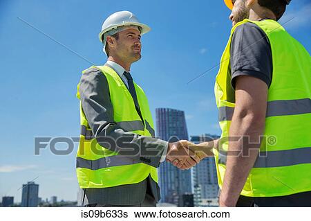 Stock Photograph - Two men wearing hard hats and hi vis vests, shaking hands, outdoors. Fotosearch