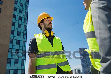 Stock Photograph - Two men wearing hard hats and hi vis vests, having conversation, outdoors. Fotosearch