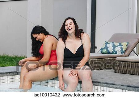 Stock Image - Two mid adult women sitting on outdoor swimming poolside laughing together, Cape Town, South Africa. Fotosearch