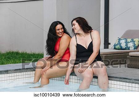 Stock Photo - Two mid adult women sitting on outdoor swimming poolside chatting and laughing together, Cape Town, South Africa. Fotosearch