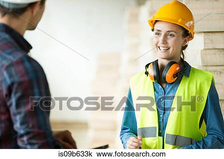 Stock Image - Two people standing in constructions site, wearing hard hats, having discussion. Fotosearch