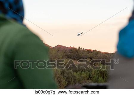 Stock Image - Two people watching helicopter above mountain landscape, over shoulder view, Cape Town, Western Cape, South Africa. Fotosearch