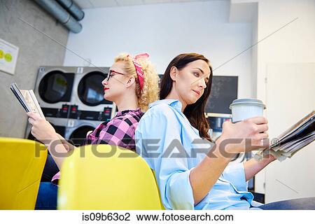 Stock Image - Two women reading newspapers back to back in laundrette. Fotosearch