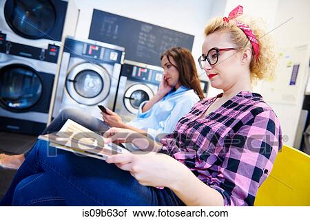 Stock Image - Two women sitting in laundrette reading magazine and looking at smartphone. Fotosearch