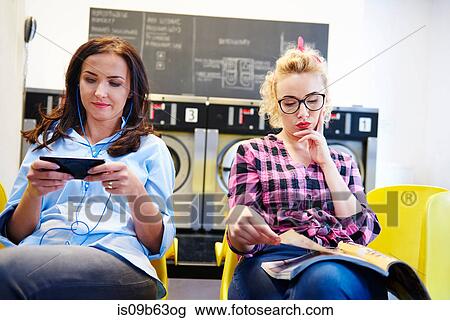 Stock Image - Two women sitting in laundrette reading magazine and looking at smartphone. Fotosearch
