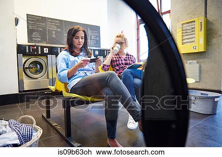 Stock Image - Two women sitting in laundrette reading magazine and looking at smartphone. Fotosearch