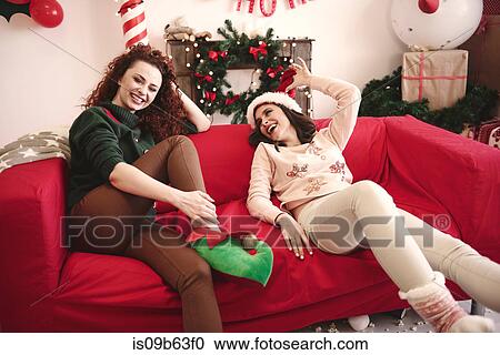 Stock Image - Two young female young friends laughing on sofa at christmas party. Fotosearch
