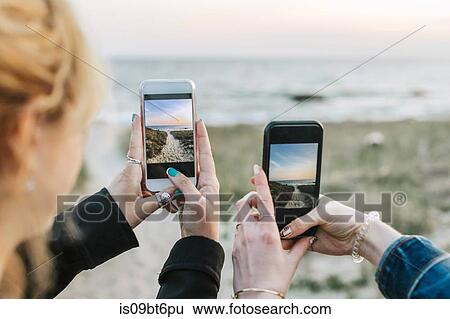 Stock Photograph - Two young women photographing beach on smartphones, over shoulder view, Menemsha, Martha's Vineyard, Massachusetts, USA. Fotosearch
