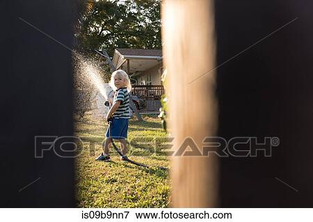Stock Photo - View through gap in fence of boy spraying water from hosepipe. Fotosearch