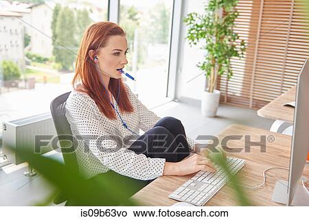 Woman at desk wearing earbuds using computer View Large Photo Image Picture - Woman at desk wearing earbuds using computer. Fotosearch